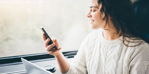 Young woman sat on train with mobile in hand & working on her laptop
