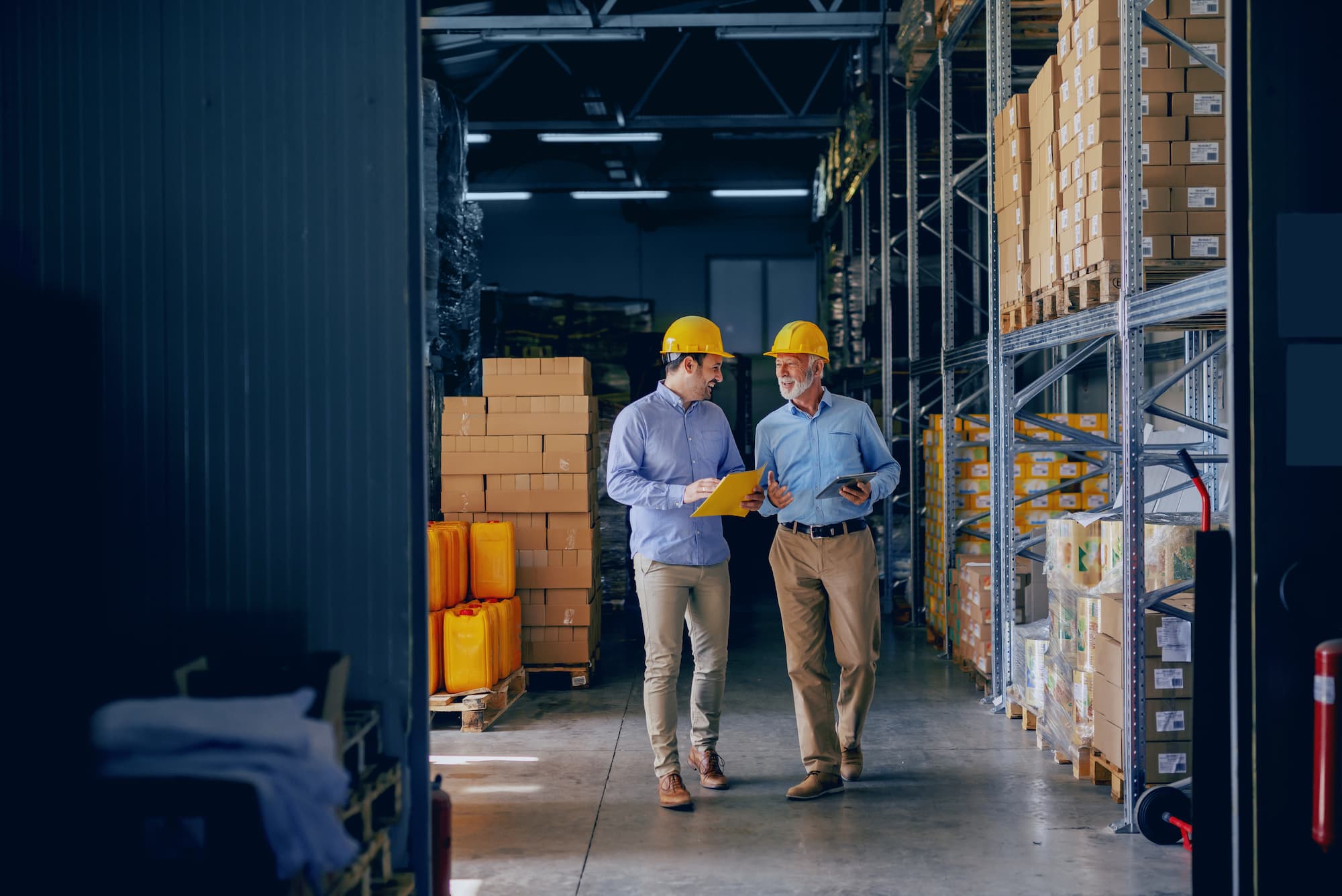 Two business partners in formal wear and with protective yellow helmets on heads walking and talking about business. Younger one holding folder with data while older one using tablet.